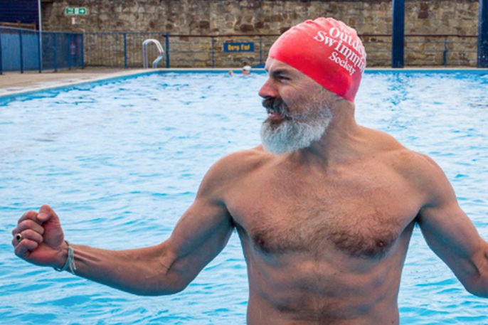 man with beard in a red swim hat with text The Outdoor Swimming Society on it in white in the waters of an outdoor pool