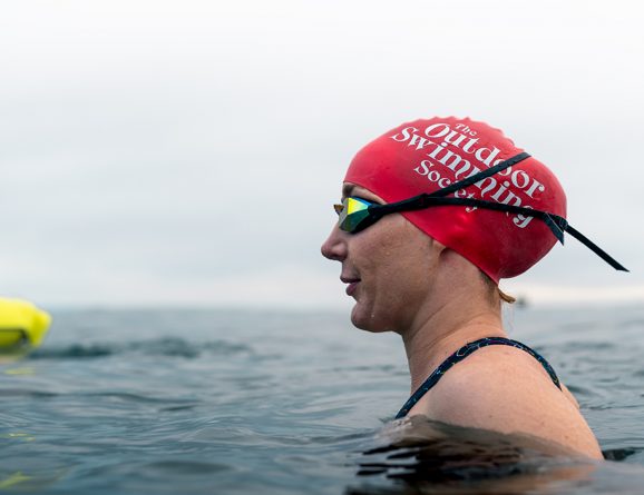 woman in water wearing large size red swim hat with text The Outdoor Swimming Society written on it in white
