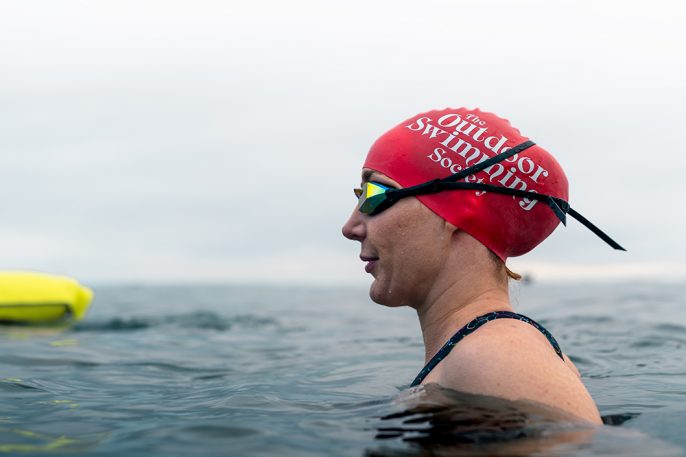 woman in water wearing large size red swim hat with text The Outdoor Swimming Society written on it in white
