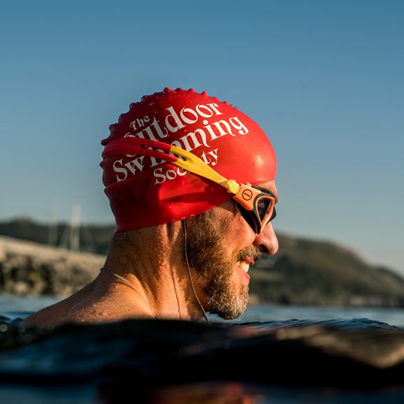 man in water wearing red swim hat with text The Outdoor Swimming Society written on it in white