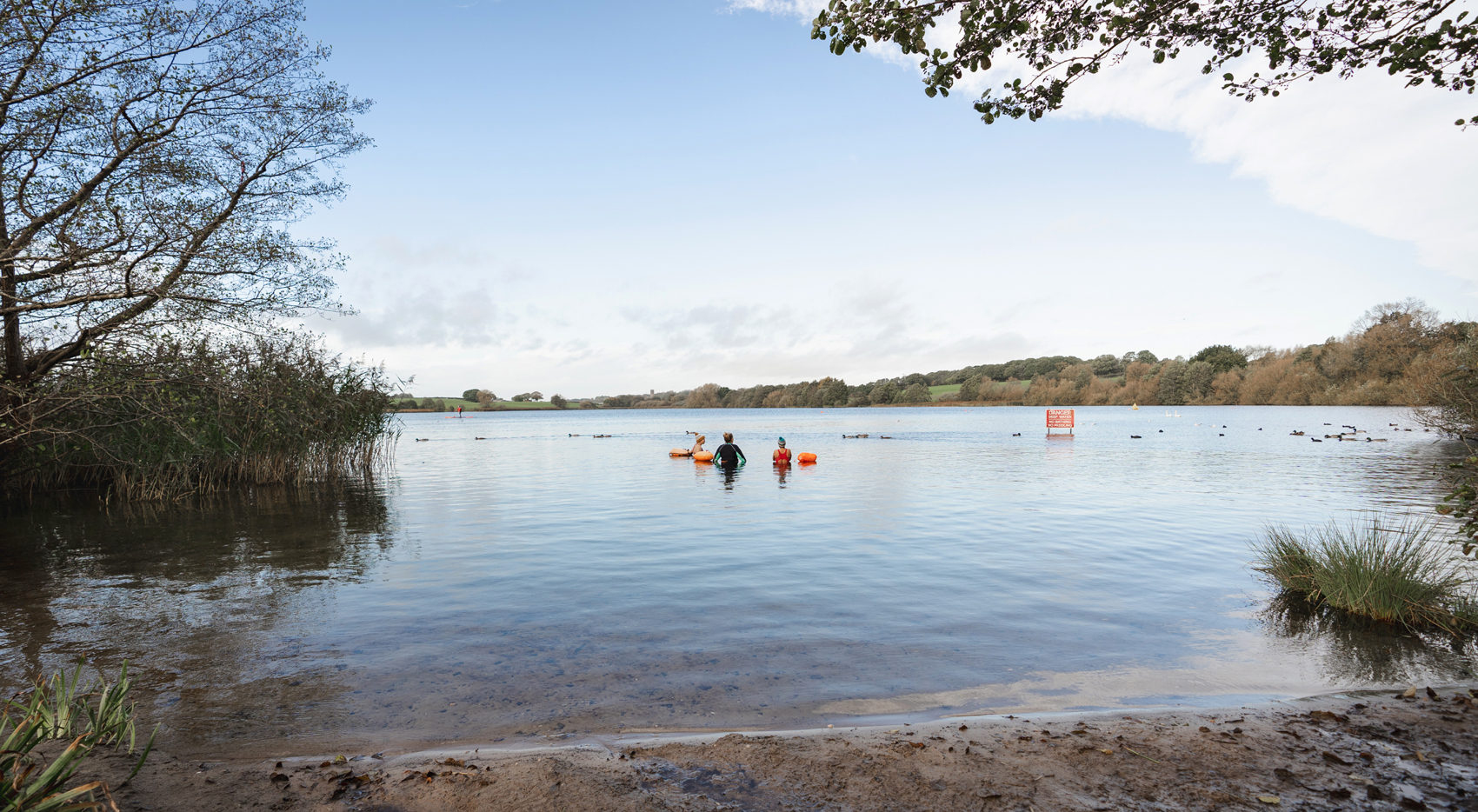 Pickmere lake framed by trees, sandy beach, still water, with three swimmers with tow floats and a Deep Water No Swimming sign