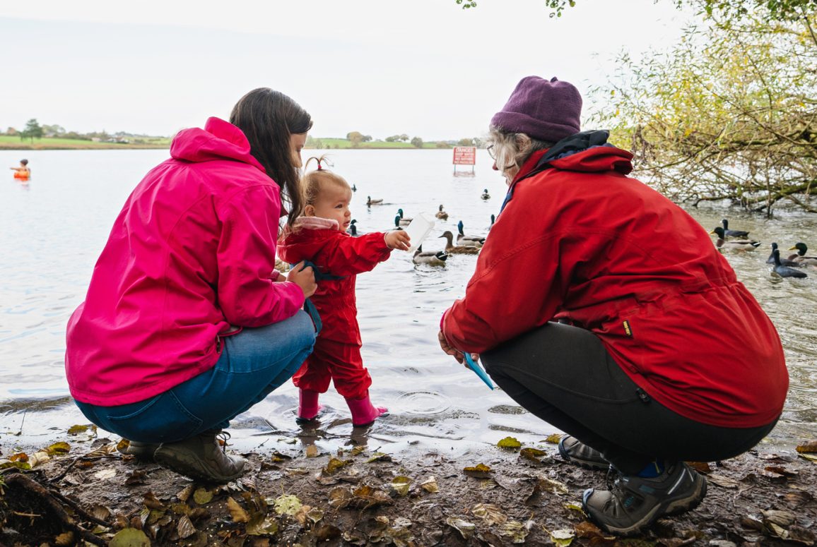 Pickmere lake, with campaigner Pam (right) and toddler grandchild looking at her and daughter (left), with ducks and 2 swimmers and No Swimming sign in background