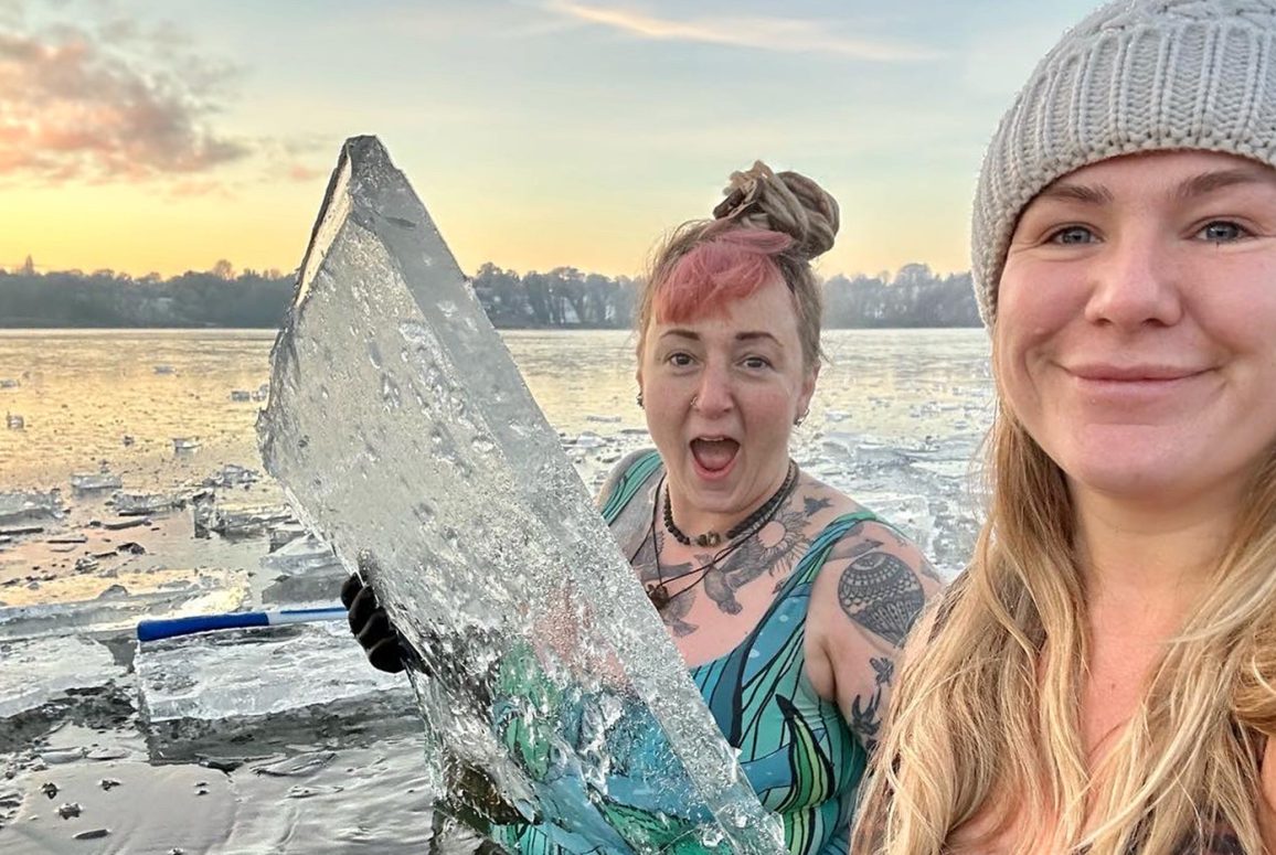 Campaigner Cat and another swimmer in swimsuits in Pickmere lake, smiling, Cat holding a large chunk of ice and grinning