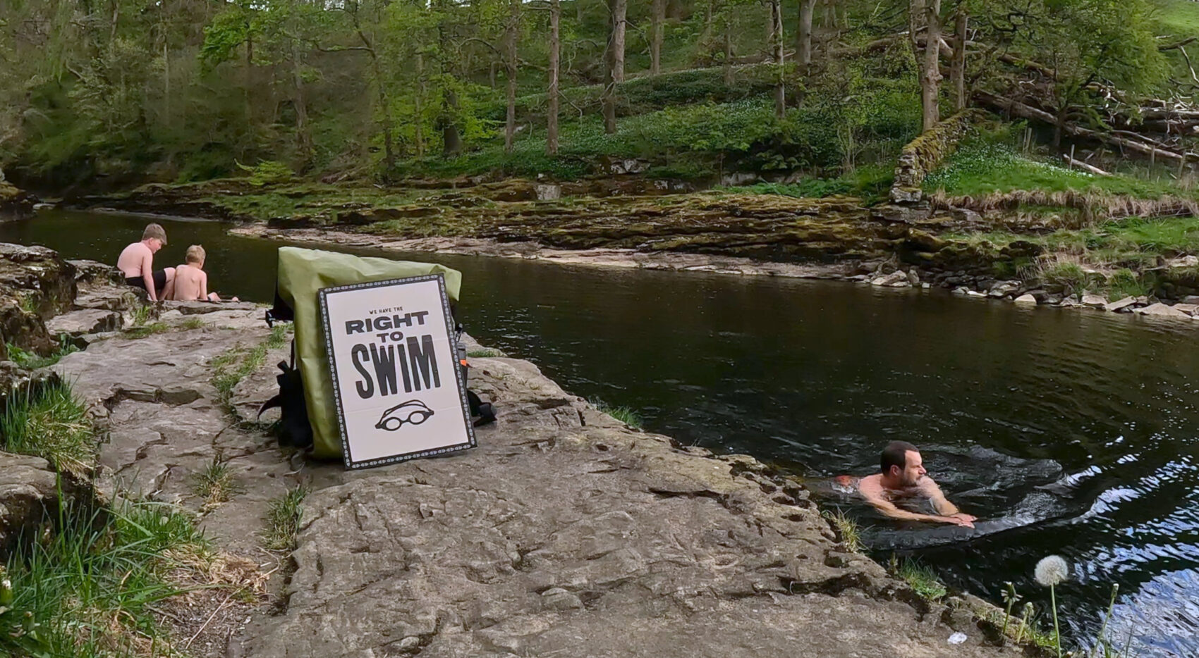 Swimmer in a river, rocky shore, wooded backdrop, 'Right to Swim placard, two children in swimming costumes on the rocks The Eel Pool, river Ribble near Settle