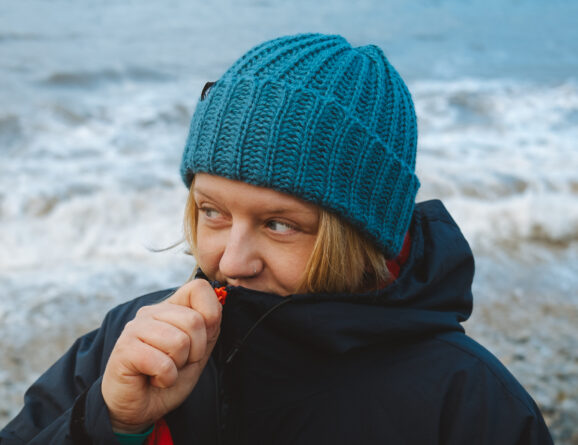 Front view of a woman in a peacock colour alpaca beanie on a pebble beach