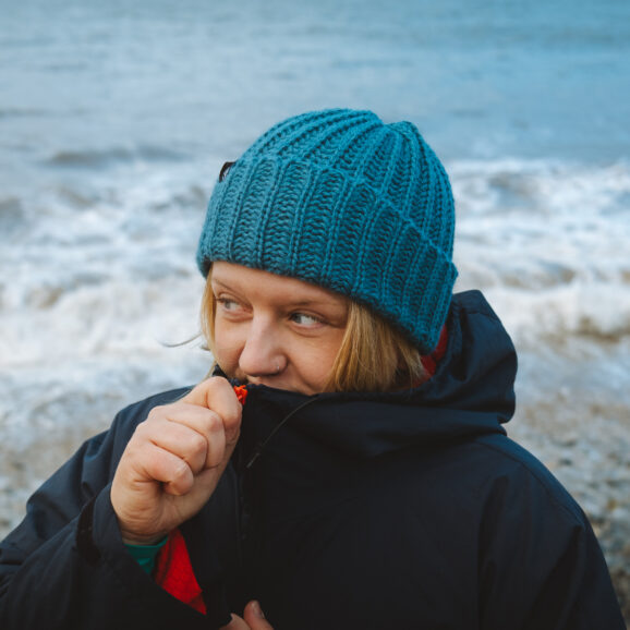 Front view of a woman in a peacock colour alpaca beanie on a pebble beach