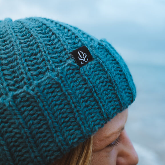 Close up side view of a woman in a peacock colour alpaca beanie on a pebble beach