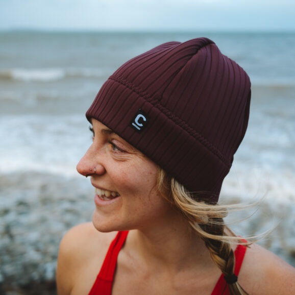 3/4 view of a woman in a rust storm chaser beanie on a pebble beach